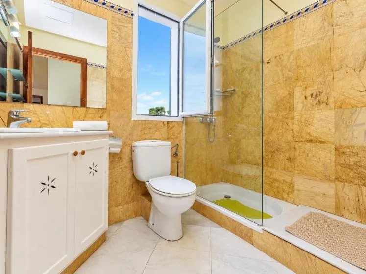 Un baño con lavabo, inodoro y ducha de vidrio junto a una ventana abierta.