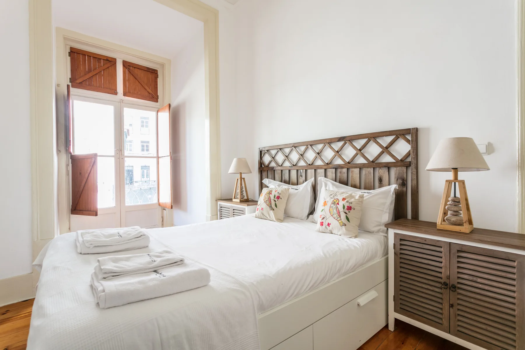A bright bedroom with a double bed, patterned pillows, and wooden shutters.