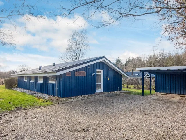 A blue cottage stands beside a gravel driveway and a small covered structure.