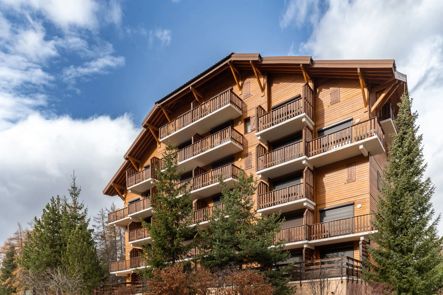 A multi-story wooden building with balconies stands among tall evergreen trees.