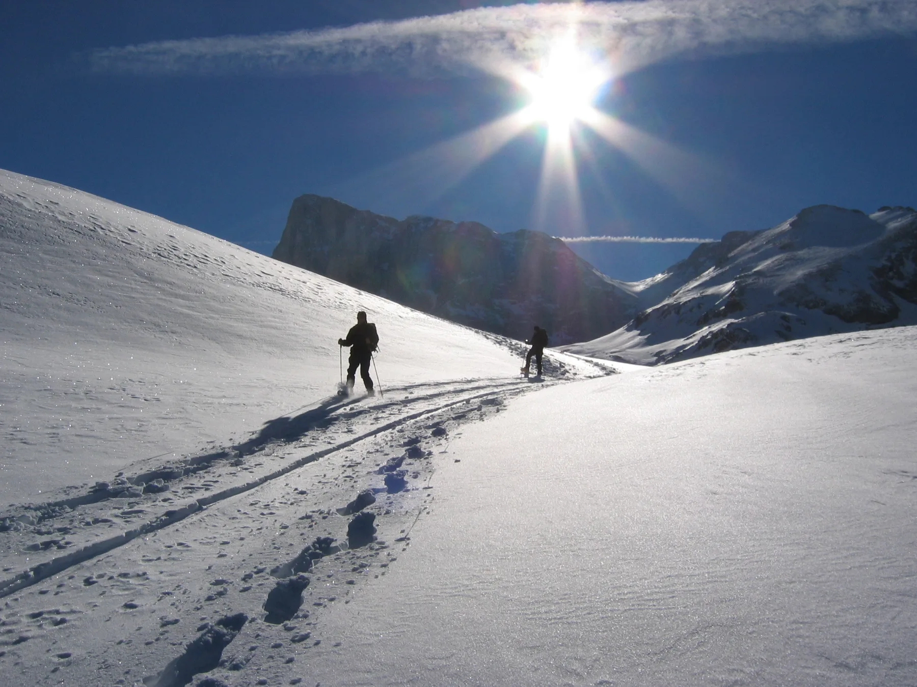 Two people ski across a sunlit snowy mountain slope.