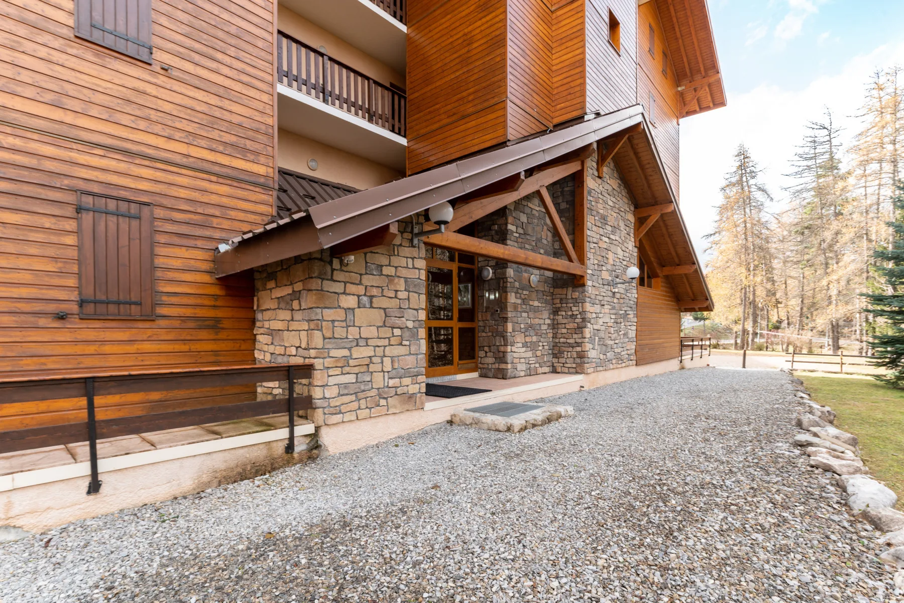 A timber-clad building with a stone entrance faces a gravel-covered outdoor area.