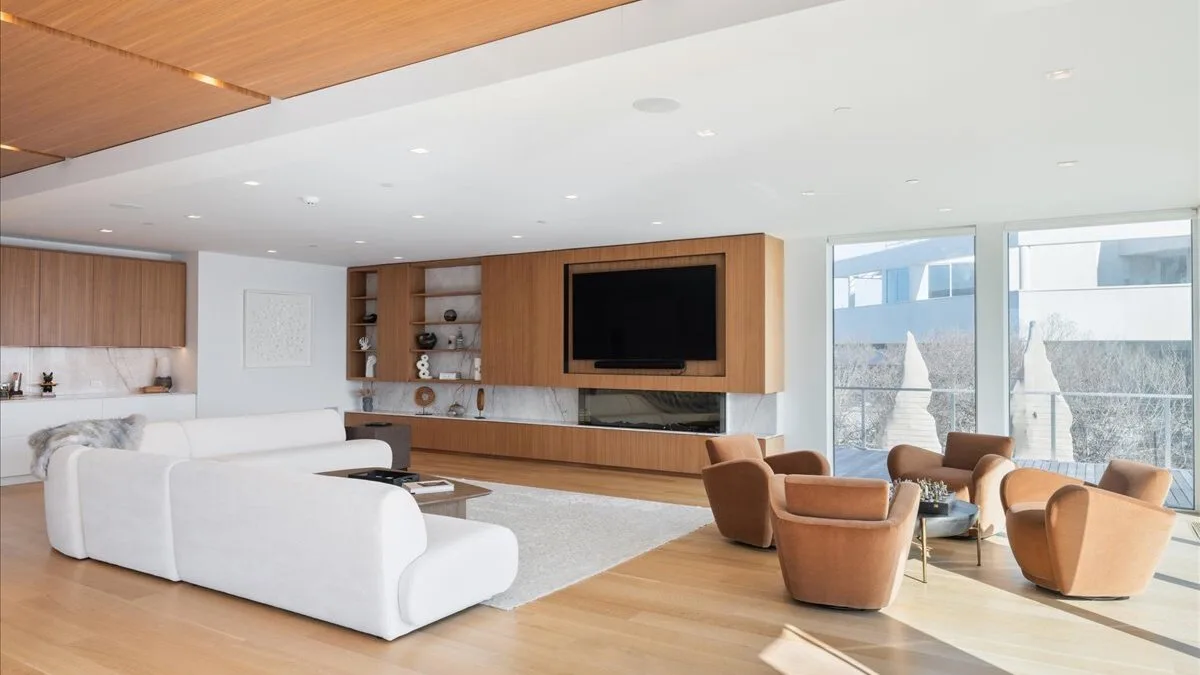 Una sala de estar moderna con asientos blancos, muebles de madera y ventanales.