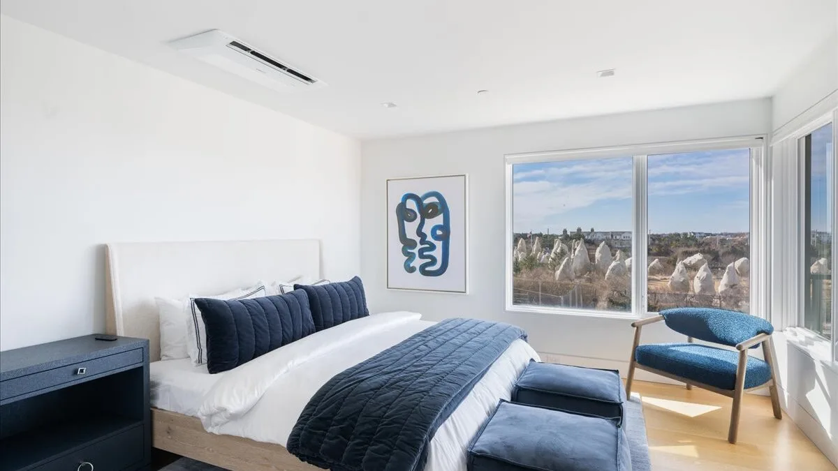 Un dormitorio luminoso con cama, ropa de cama azul y grandes ventanas con vistas a la ciudad.
