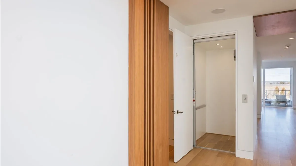 Un pasillo interior luminoso con una puerta abierta y una habitación con suelo de madera al fondo.