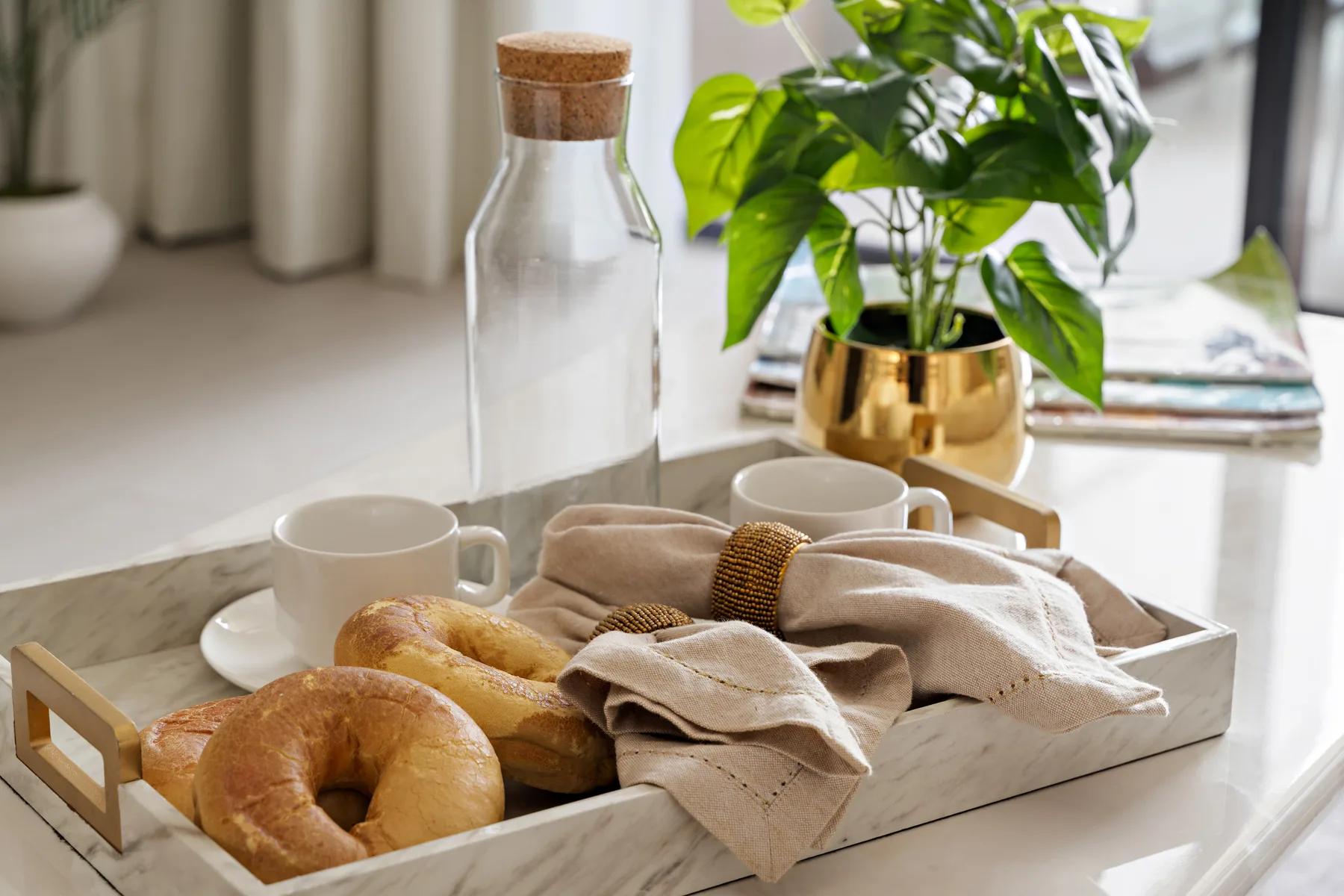 A breakfast tray with pastries and cups sits beside a glass bottle and a leafy plant.