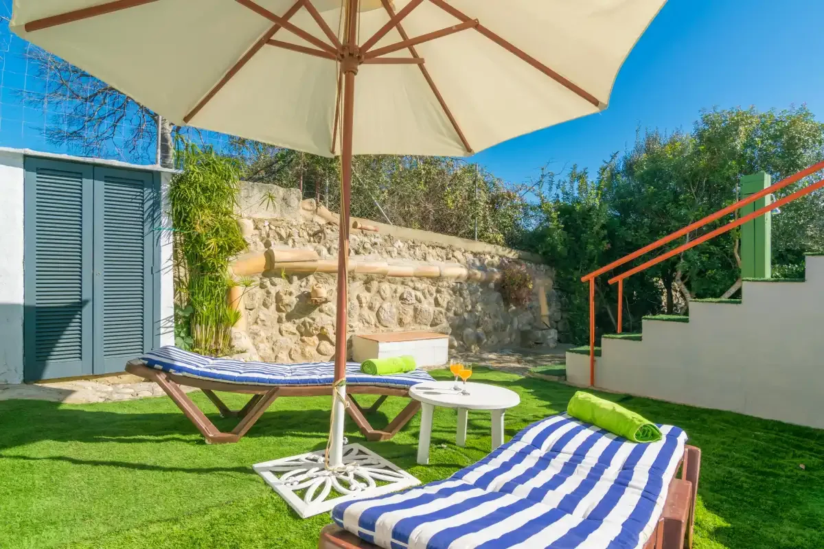 A sunny outdoor seating area has striped loungers, a table, and a large umbrella.