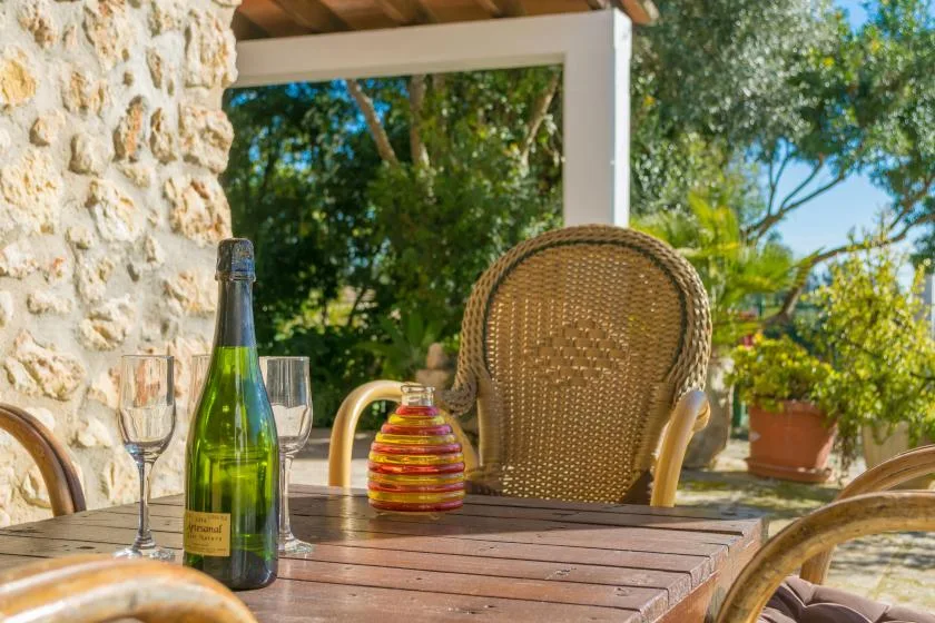 An outdoor dining table with a bottle, glasses, and woven seating under a shaded patio.