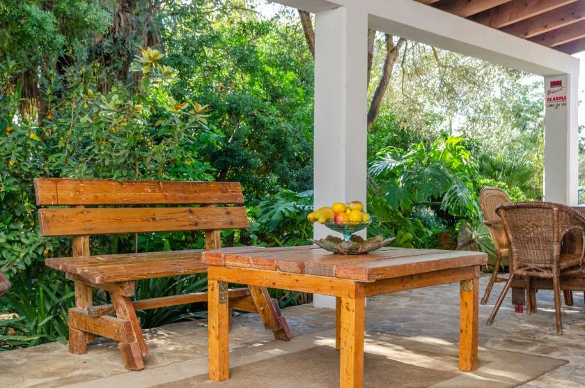 A covered outdoor patio with wooden seating and a table surrounded by greenery.