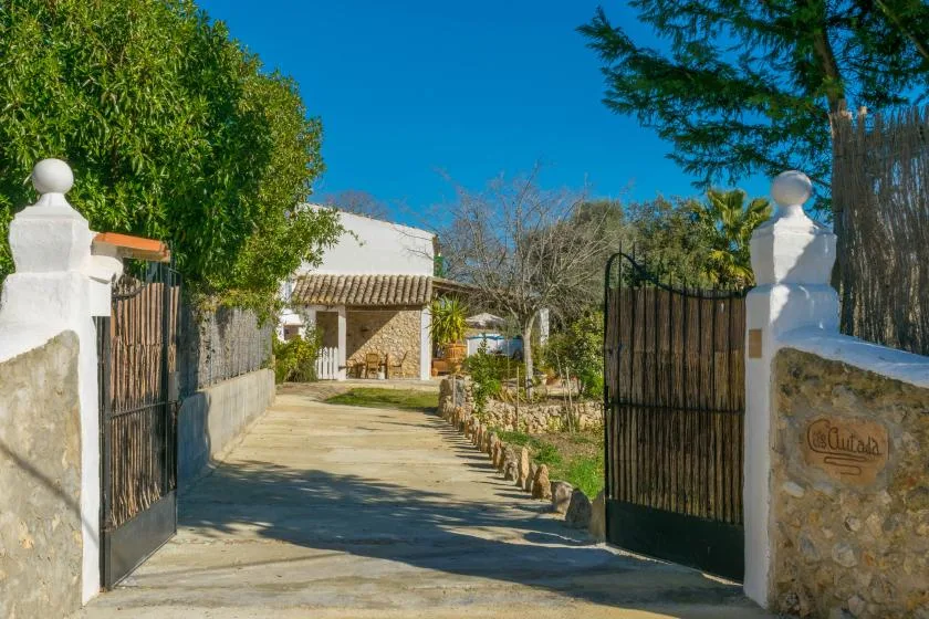 A paved driveway passes through an open gate toward a building and garden.