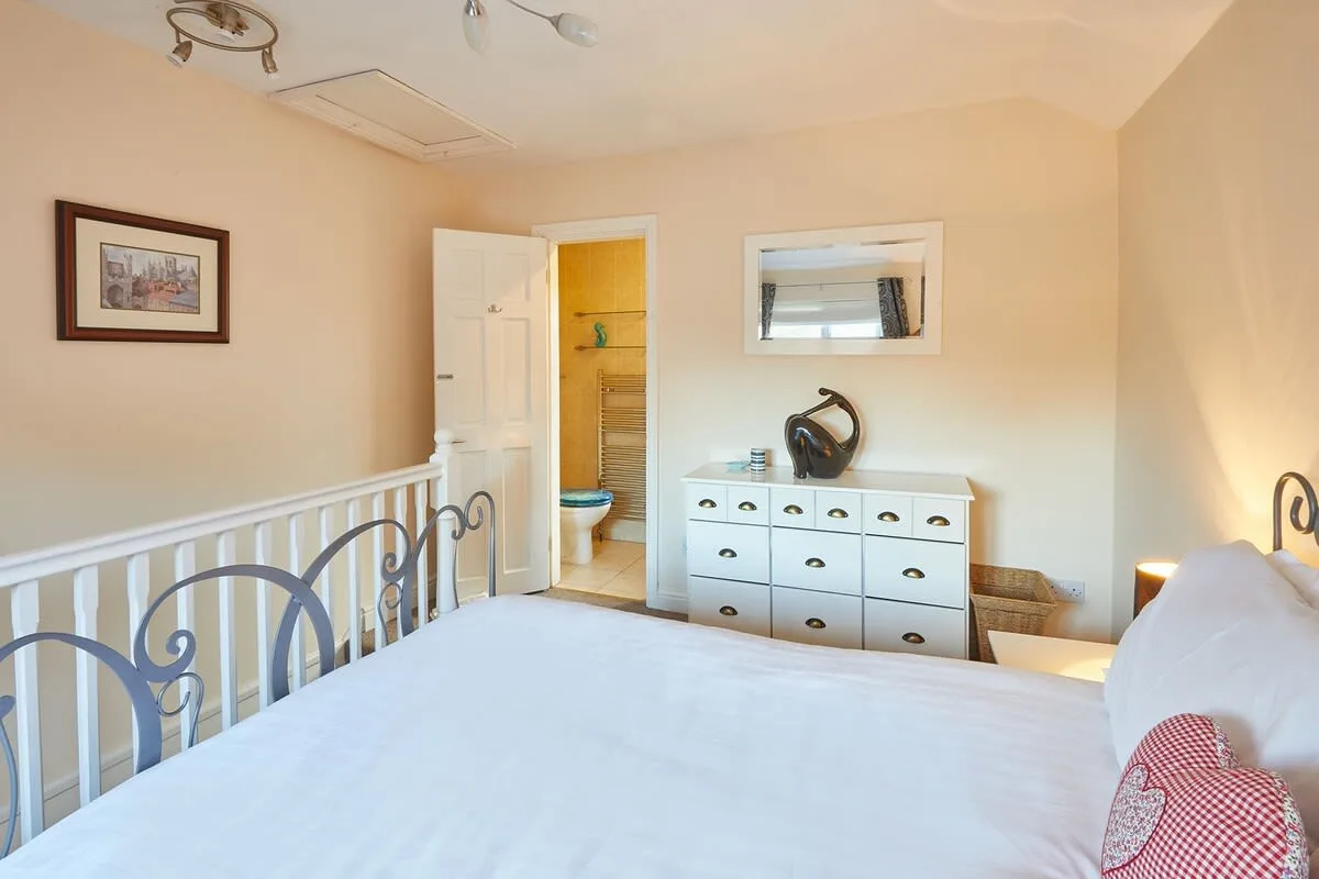 Dormitorio con cama blanca, cómoda y una puerta abierta hacia el baño.