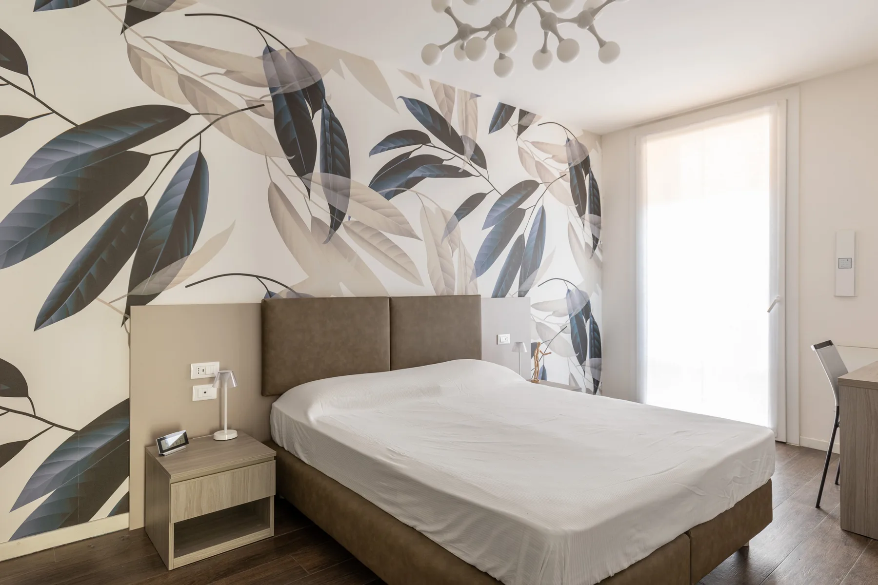 A bedroom with a bed, patterned botanical wallpaper, and a bright window.
