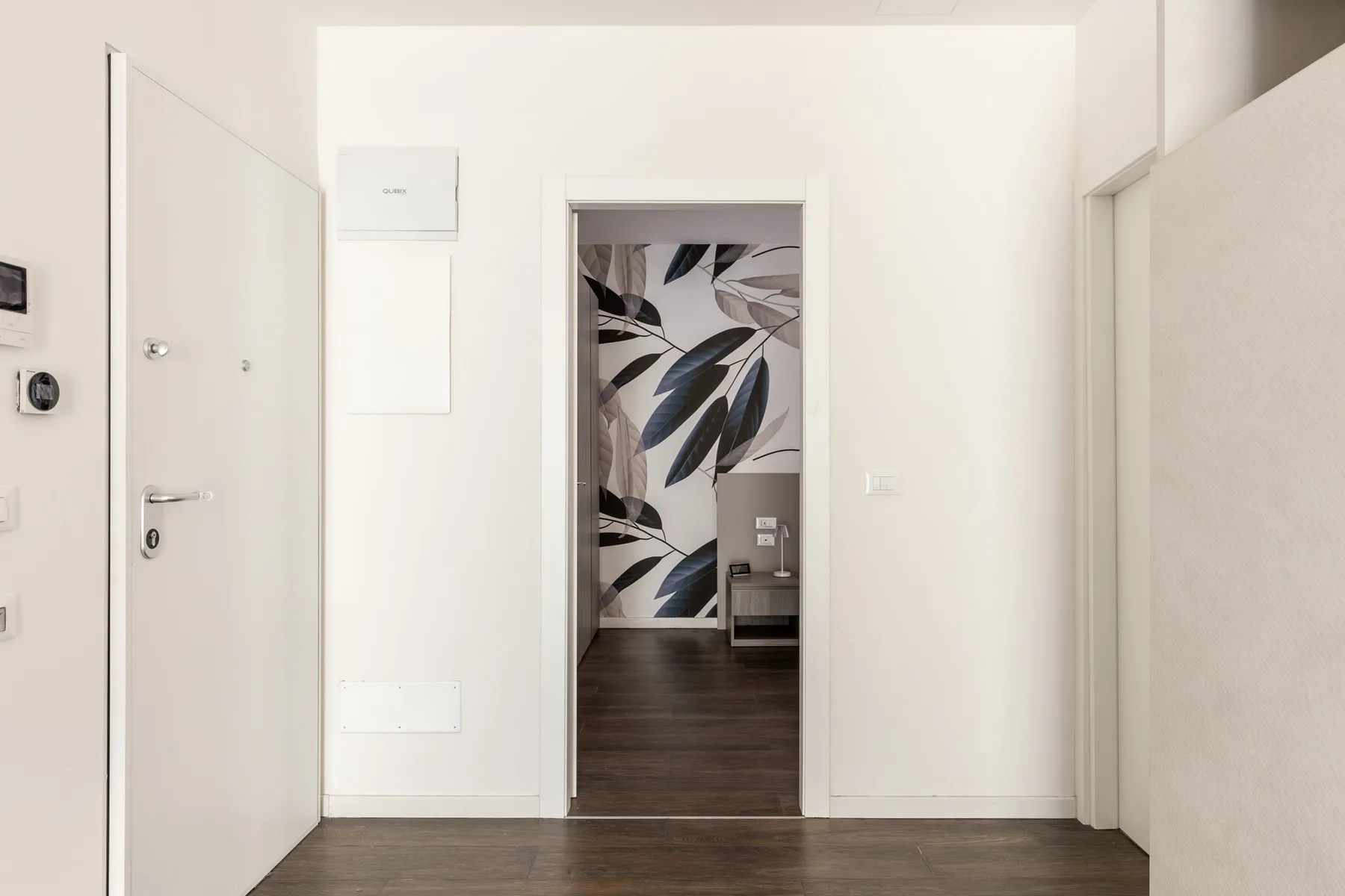 Un pasillo luminoso da a una habitación con suelo oscuro y una pared estampada.
