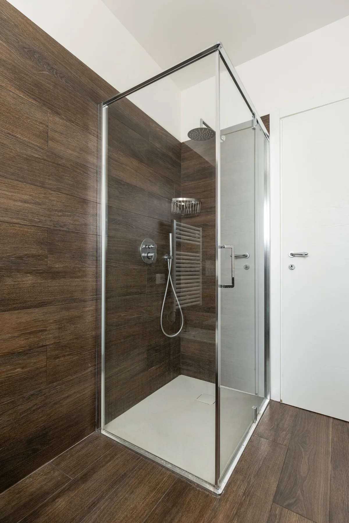 Una ducha de vidrio está junto a una puerta blanca en un baño con paredes de aspecto de madera.