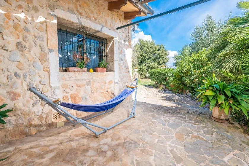 Un patio de piedra tiene una hamaca azul junto a una ventana enrejada y plantas frondosas.