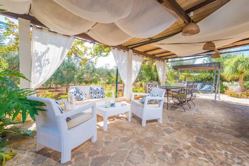 Un patio cubierto tiene asientos blancos, cortinas translúcidas y una mesa de comedor.