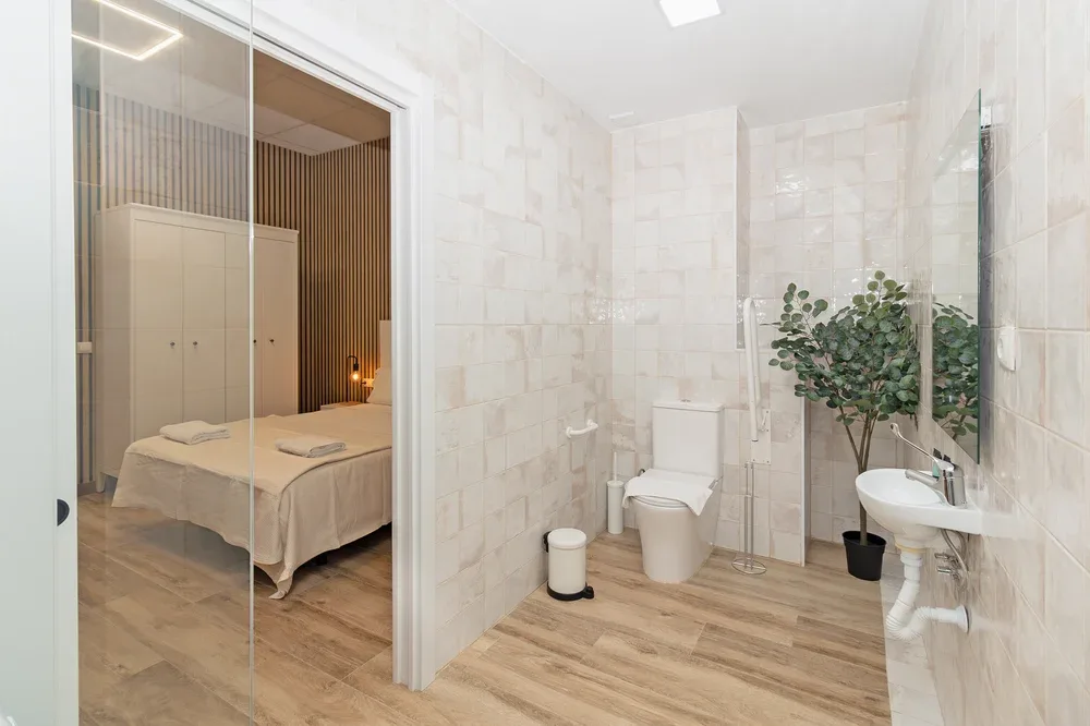 A tiled bathroom with a sink and toilet, beside a bedroom visible through a glass door.