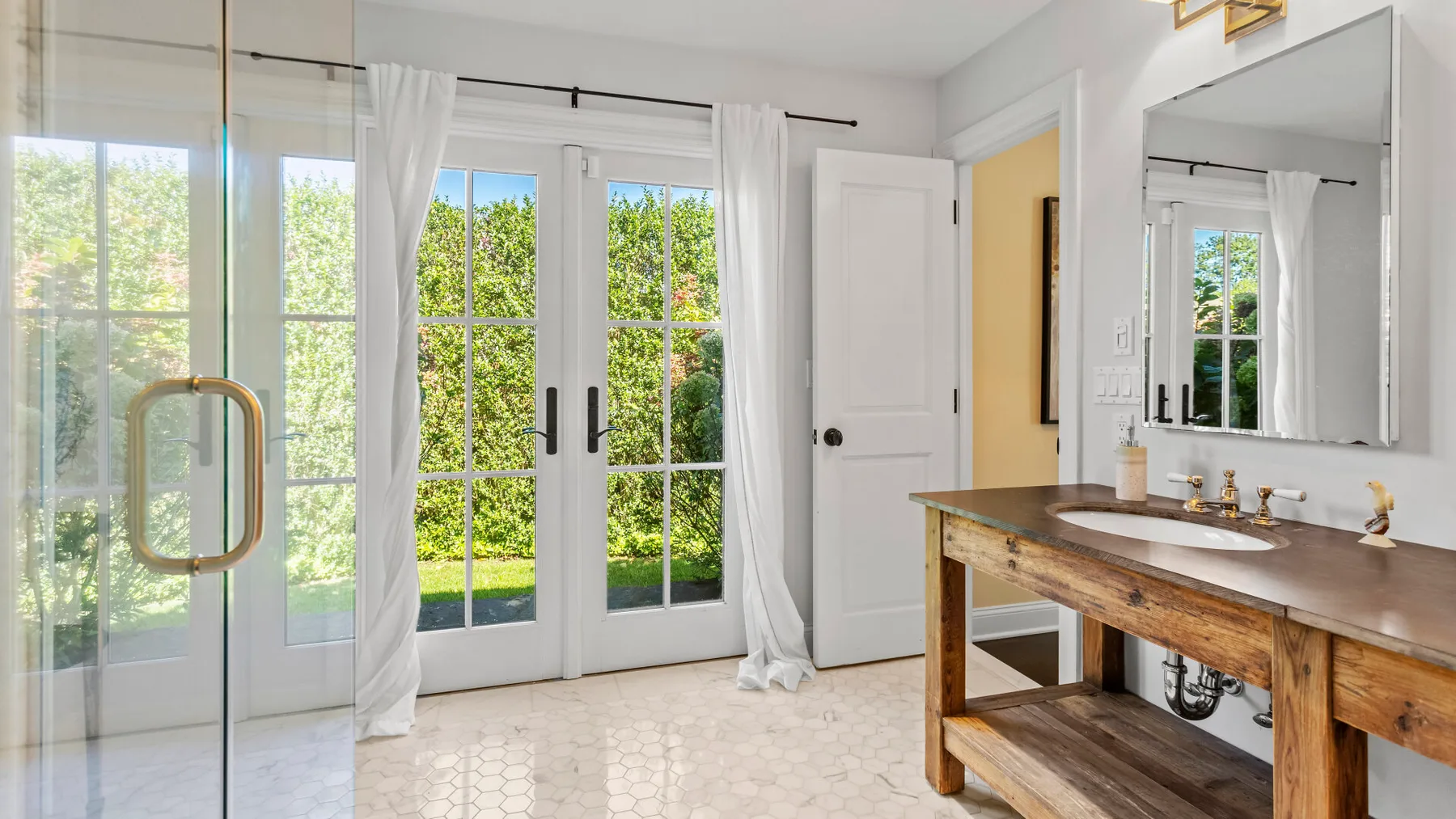 Un baño luminoso con un tocador de madera y puertas de vidrio que dan al jardín.