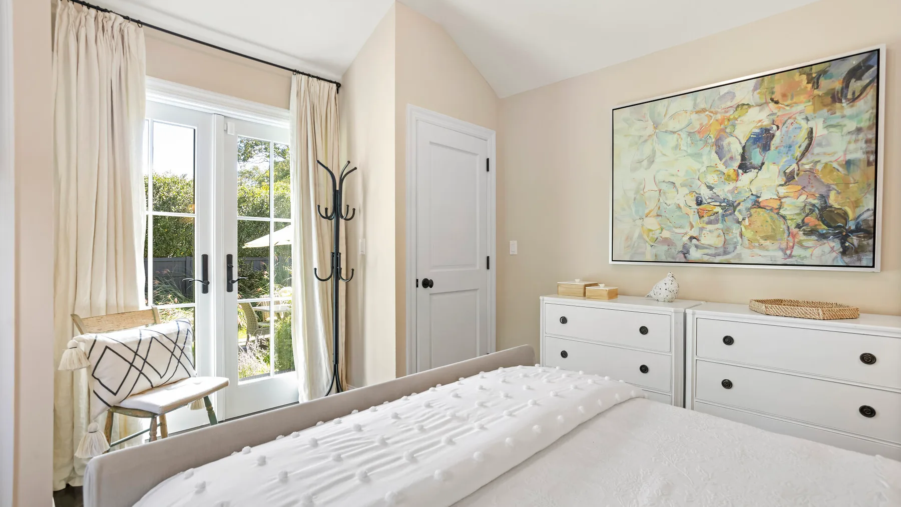 Un dormitorio luminoso con una cama, cómodas blancas y un cuadro grande y colorido.
