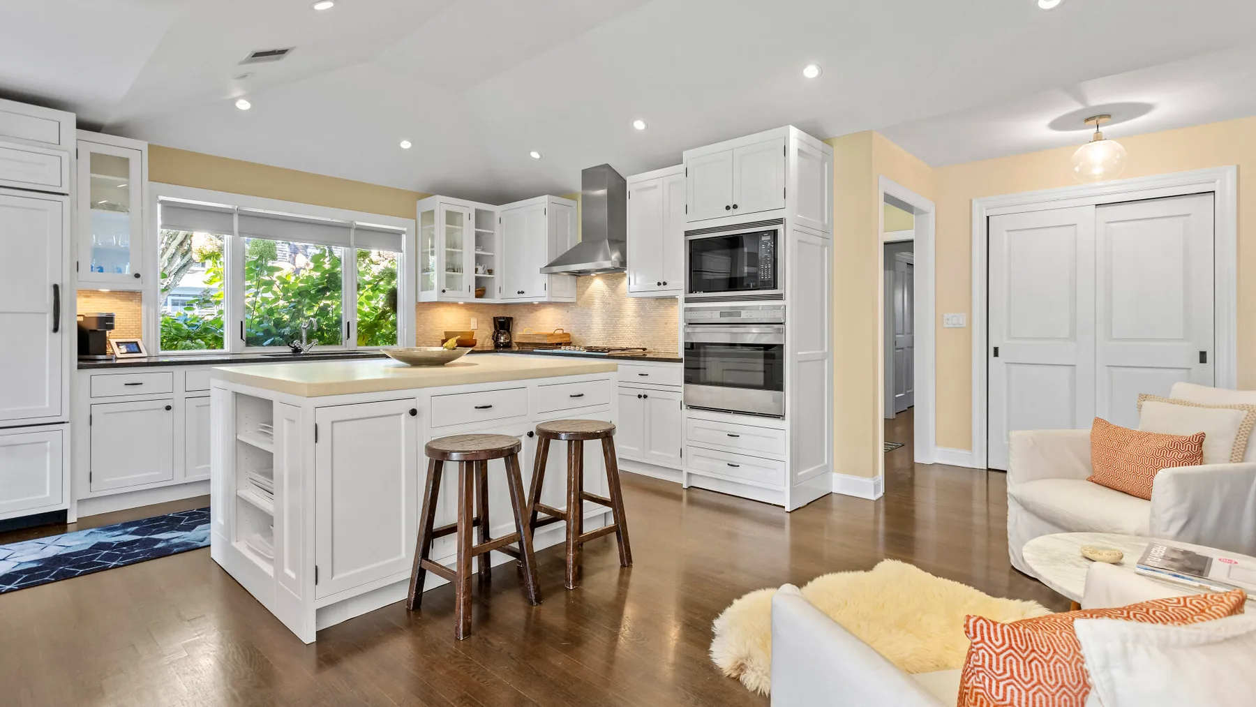 Una cocina luminosa con gabinetes blancos e isla se abre a una zona de estar.