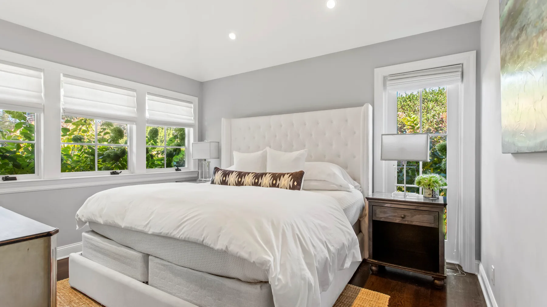 Un dormitorio luminoso cuenta con una cama grande junto a ventanas y una mesita de madera.