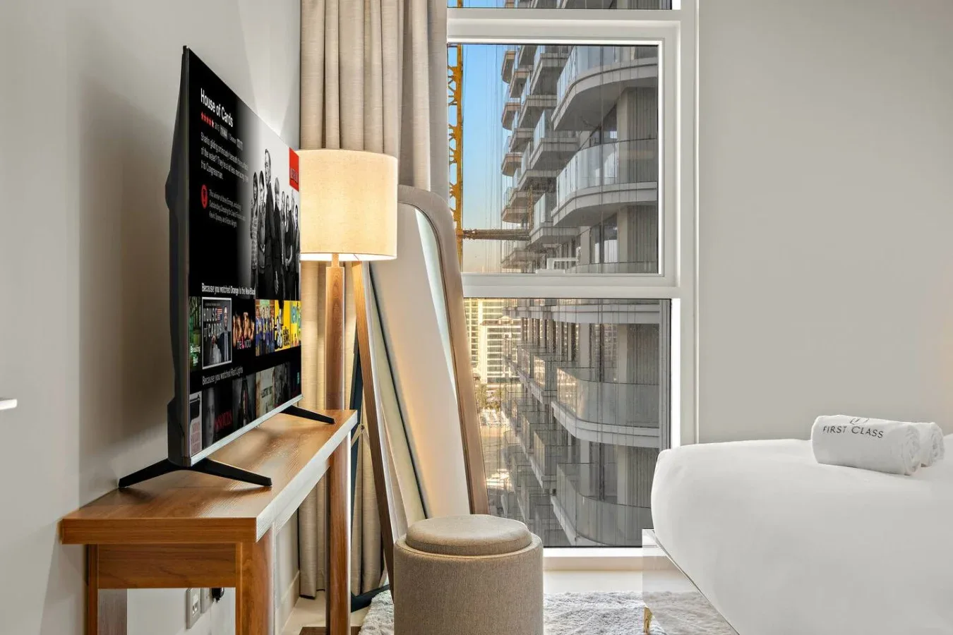 Una sala luminosa tiene un televisor, asientos y una ventana grande con vista a edificios cercanos.