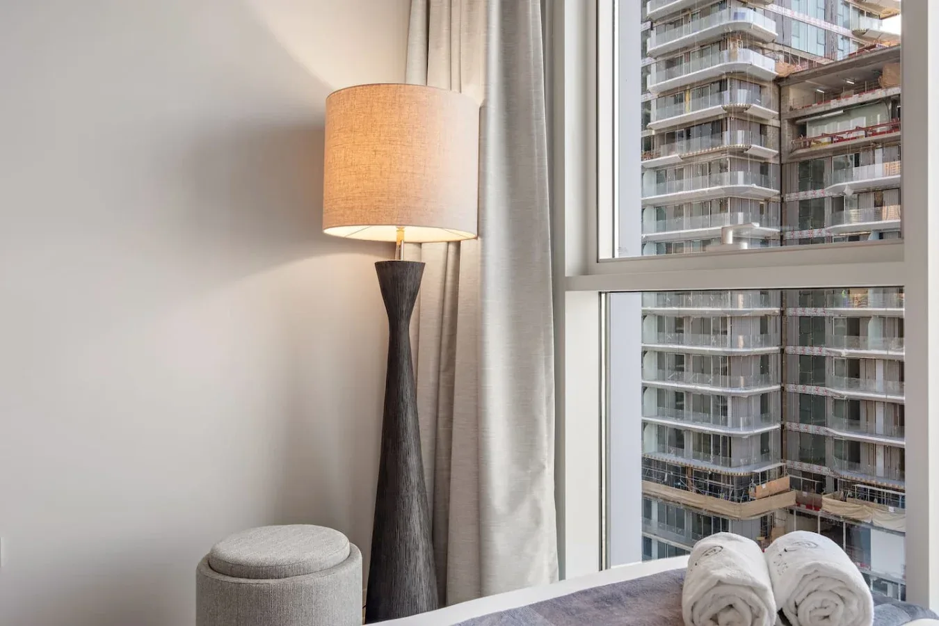 Un rincón de dormitorio con una lámpara de pie junto a una ventana con edificios a la vista.