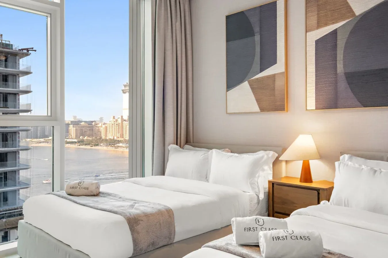 Un dormitorio luminoso con ropa de cama blanca, arte abstracto y grandes ventanas con vistas al agua.