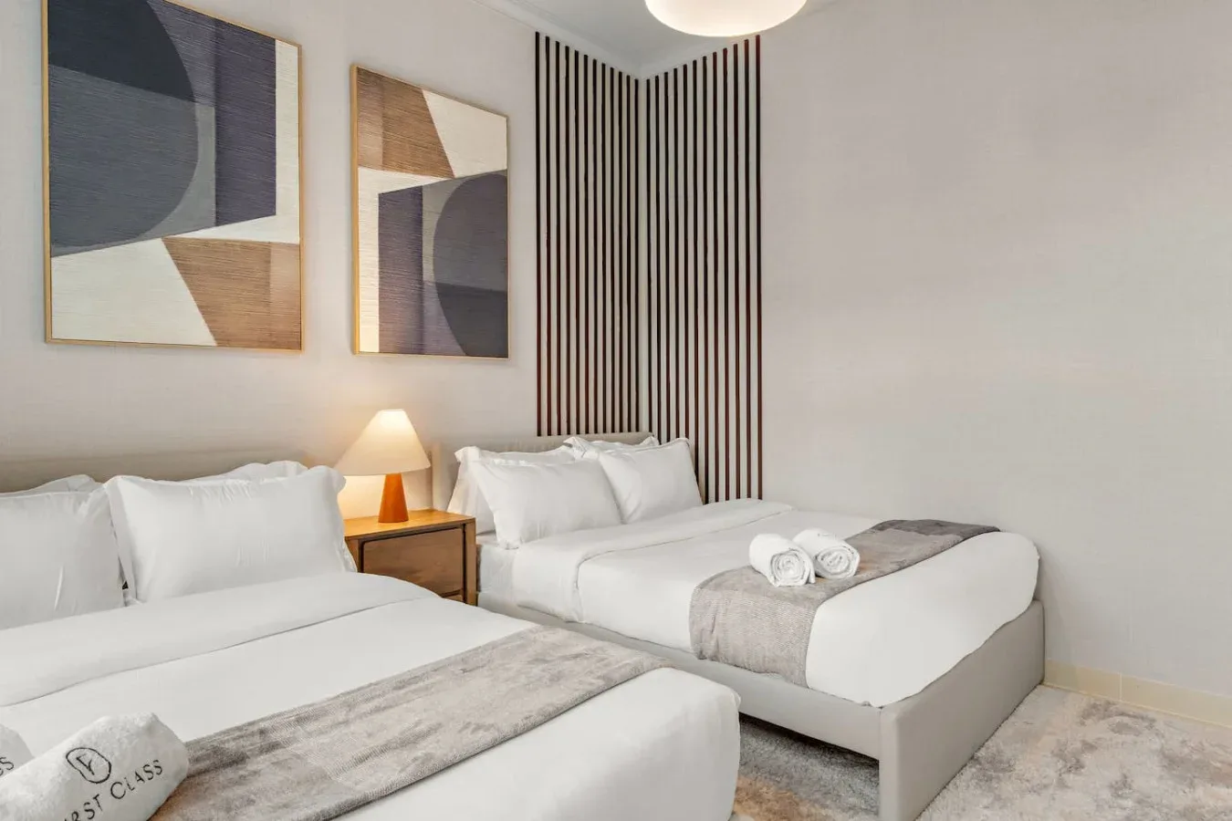 Un dormitorio luminoso con dos camas, ropa de cama neutra y arte abstracto en la pared.