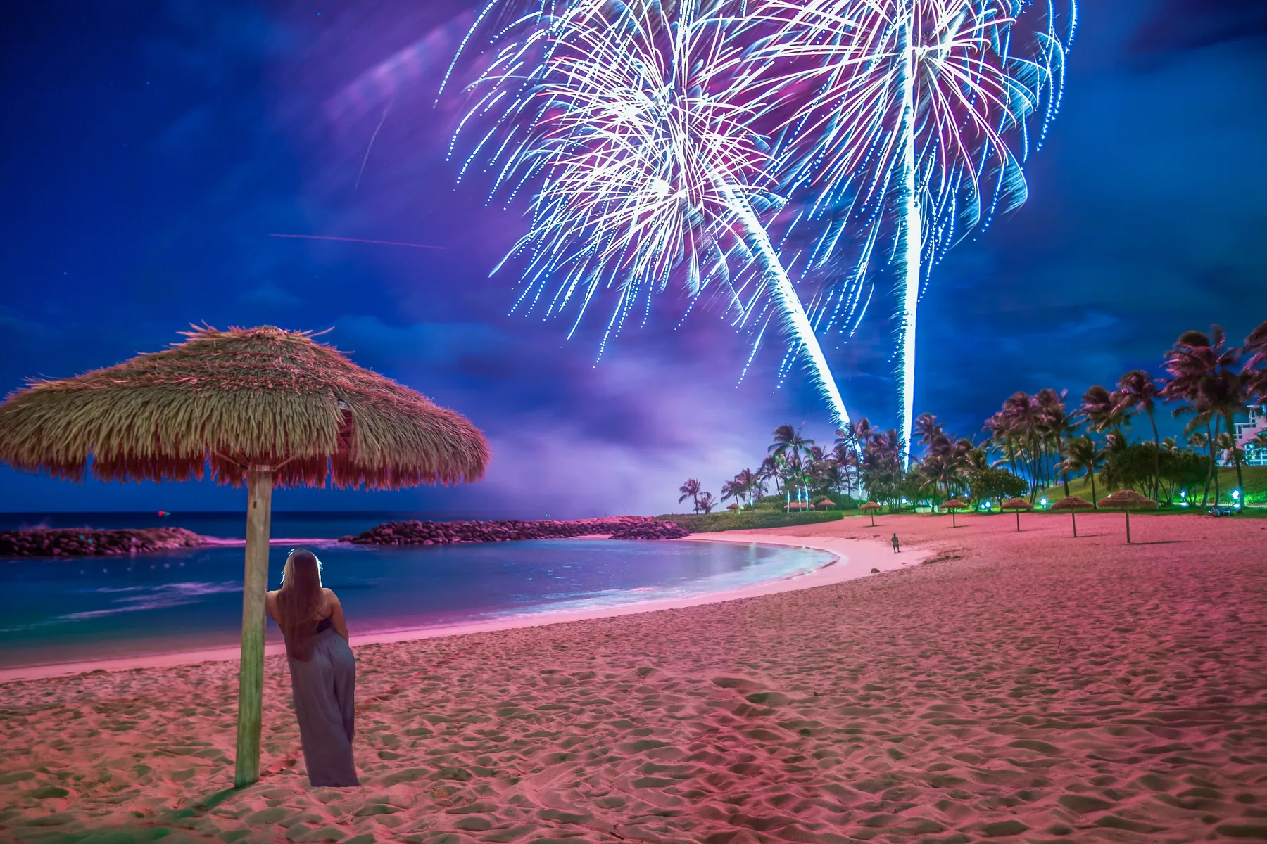 Una playa de noche con fuegos artificiales sobre el agua y una sombrilla de techo de paja.