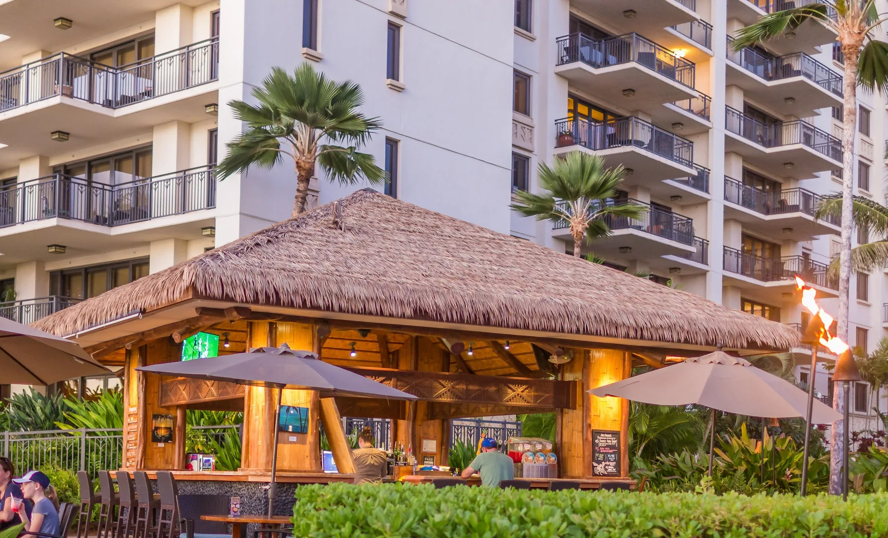 Un restaurante al aire libre con techo de paja se encuentra frente a edificios y palmeras.