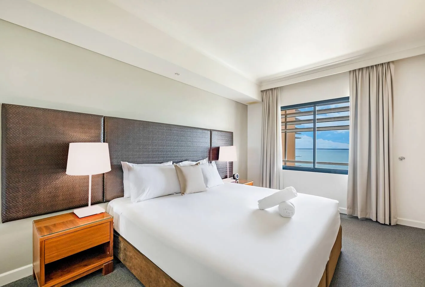 A bedroom with a neatly made bed beside a window overlooking the ocean.