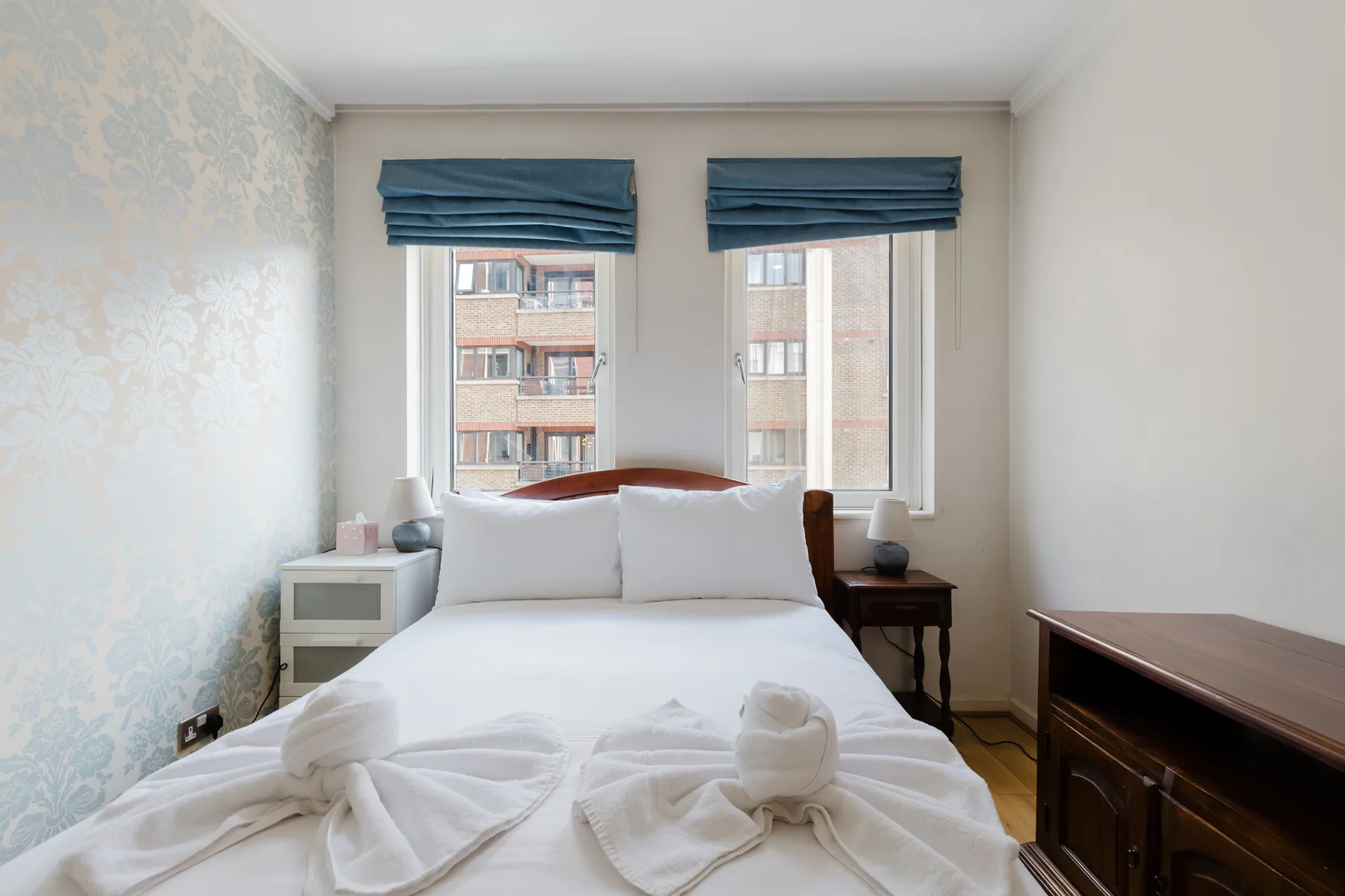 Un dormitorio con una cama bien tendida bajo dos ventanas con cortinas azules.