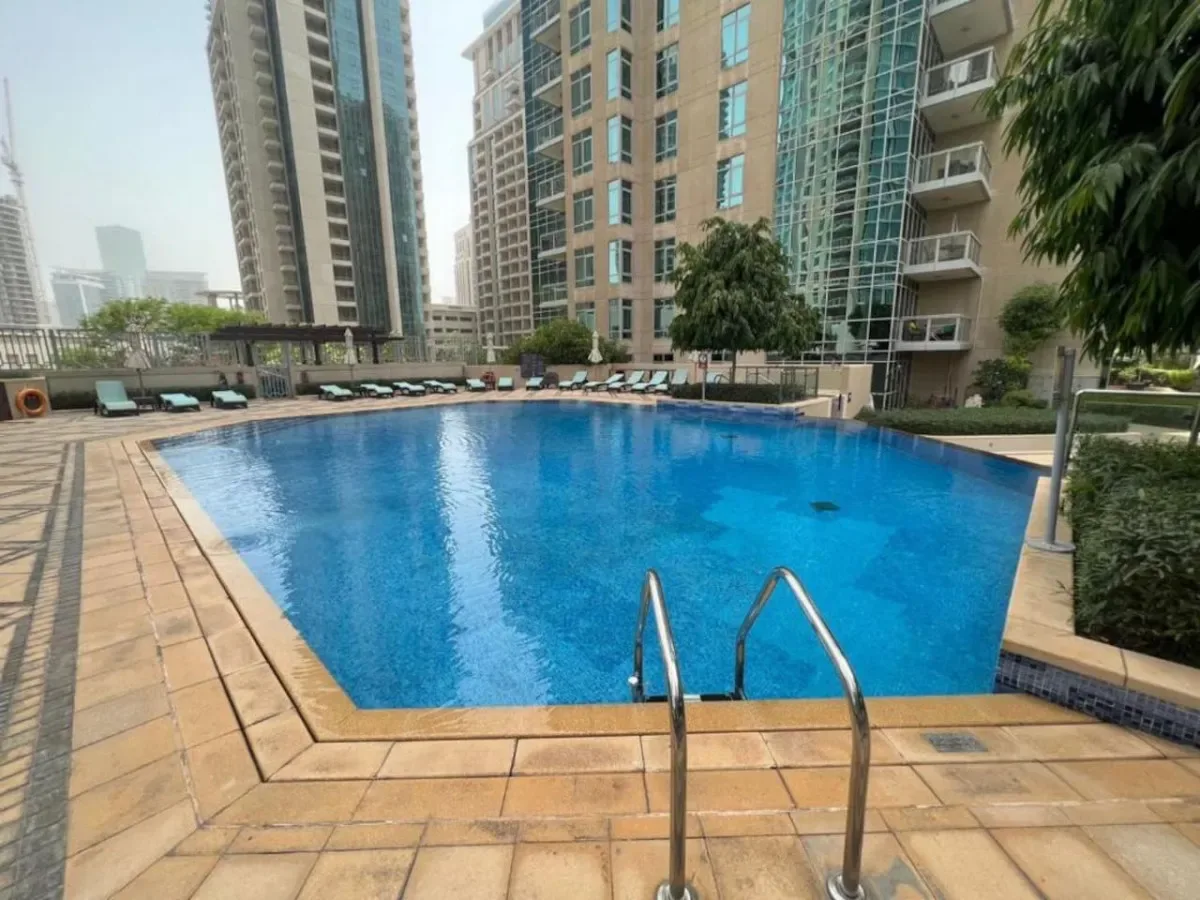 An outdoor swimming pool is surrounded by paved decking and tall buildings.
