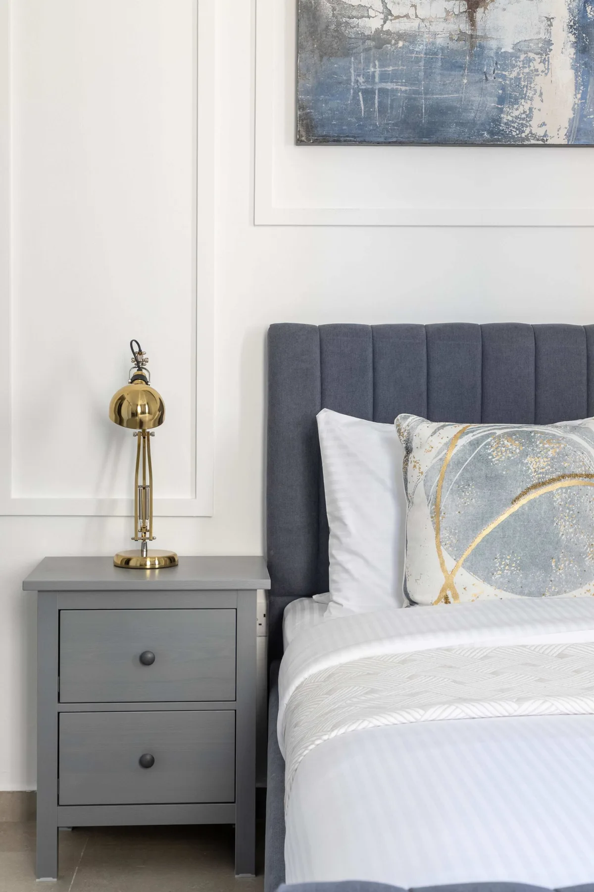 Un dormitorio con una cama gris, ropa de cama blanca y una mesita de noche.
