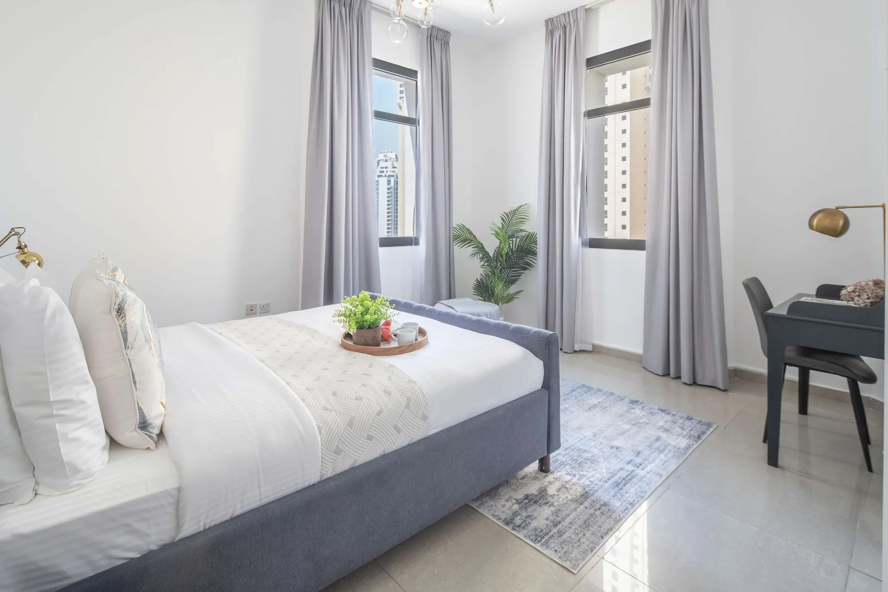 Un dormitorio luminoso con una cama bien tendida, ventanas altas y cortinas grises.