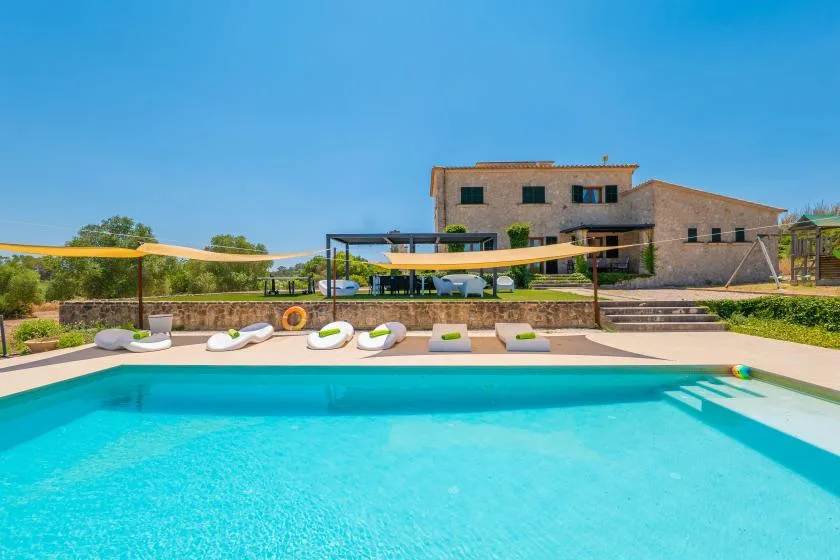 An outdoor swimming pool sits in front of a modern house and shaded loungers.