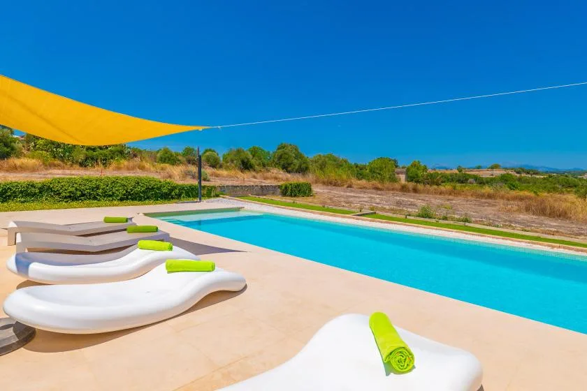 An outdoor swimming pool is bordered by loungers beneath a yellow shade sail.