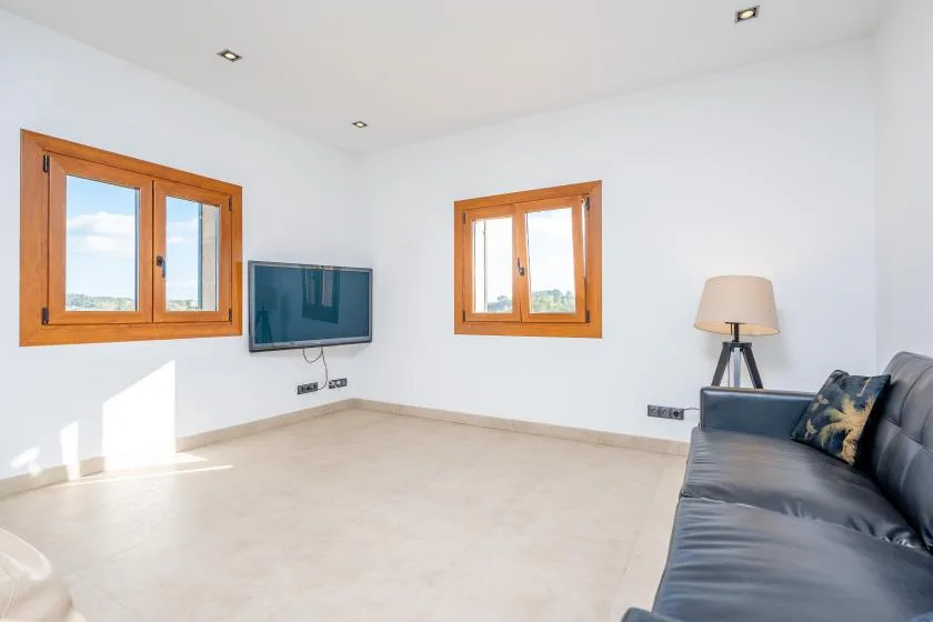 A bright living room with a wall-mounted television, wooden-framed windows, and seating.