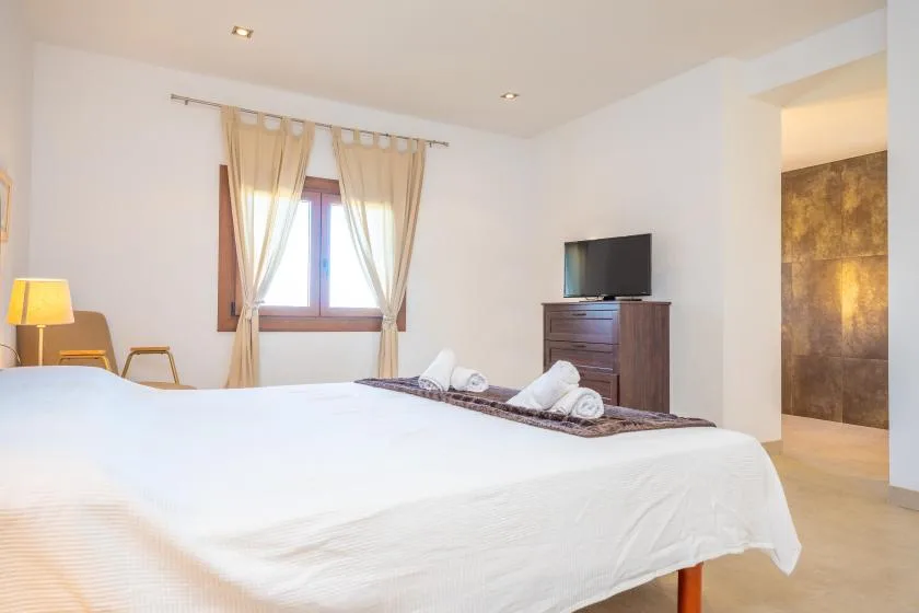 A bright bedroom with a large bed, curtained window, and television.