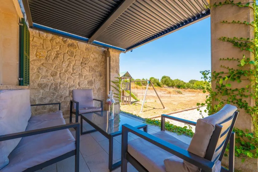A covered patio with cushioned seating overlooks a sunny open landscape.