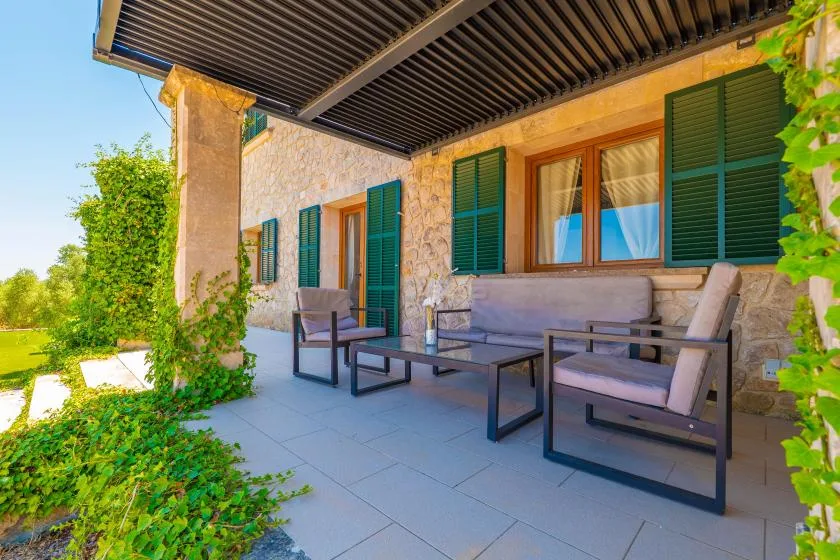 A covered stone patio with outdoor seating and green shutters.