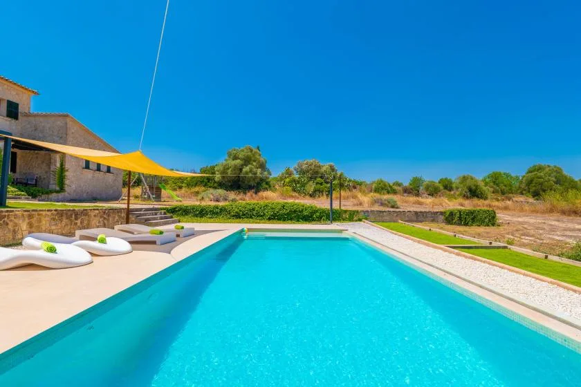 An outdoor swimming pool beside a sunlit patio with loungers and a shade canopy.
