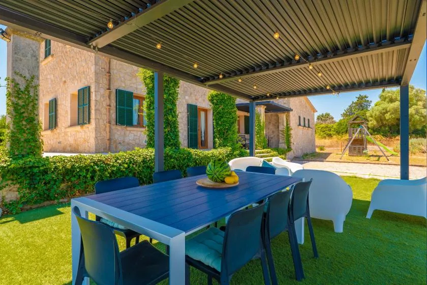 A shaded outdoor dining area beside a stone house, with a table and chairs.