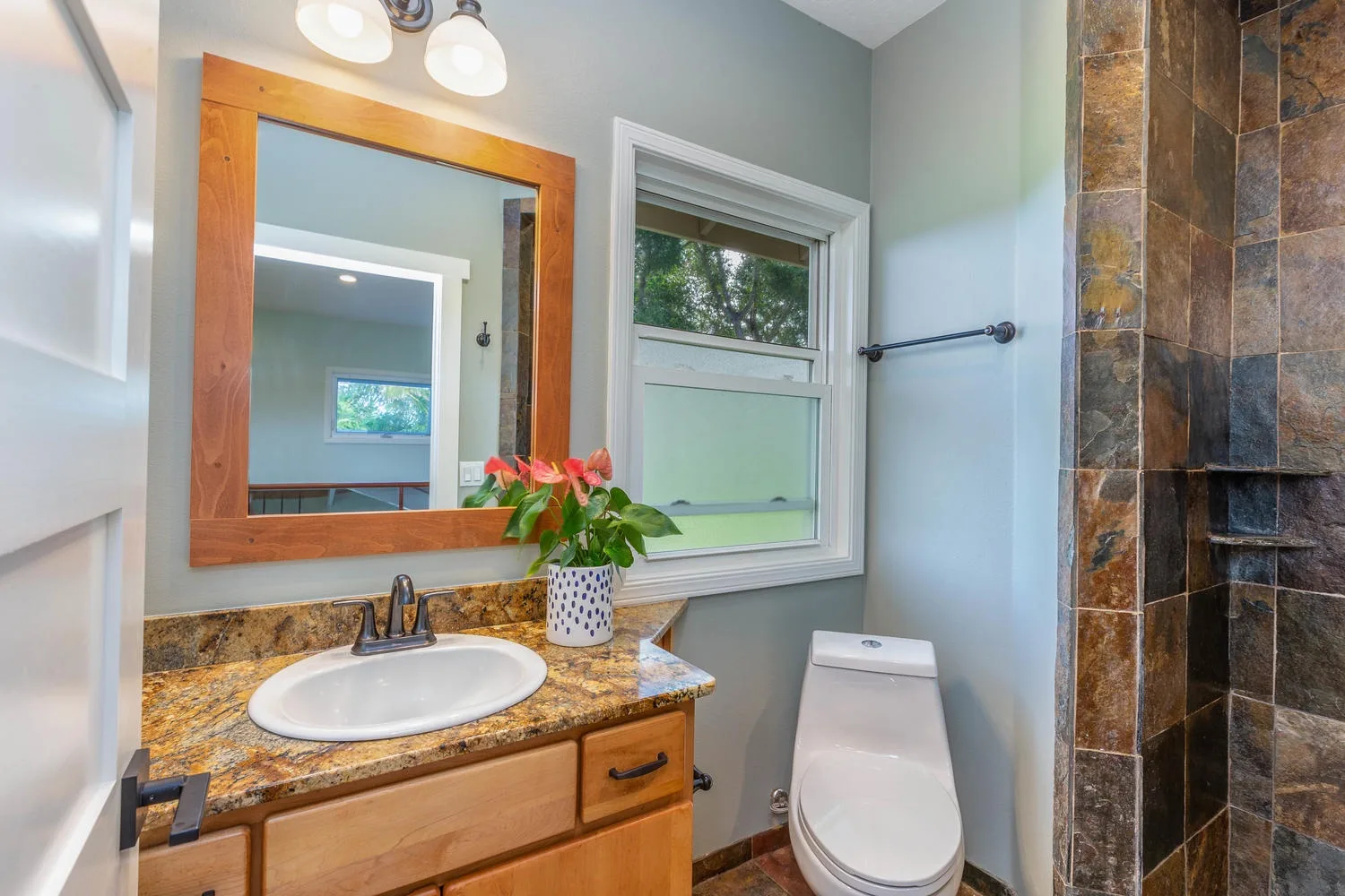 A bathroom with a vanity, mirror, window, toilet, and tiled shower.
