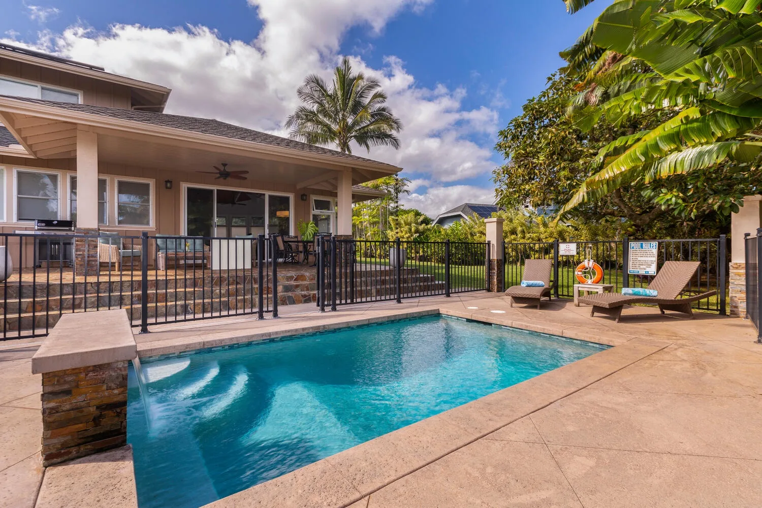 An outdoor swimming pool beside a covered patio, with lounge seating and tropical greenery.