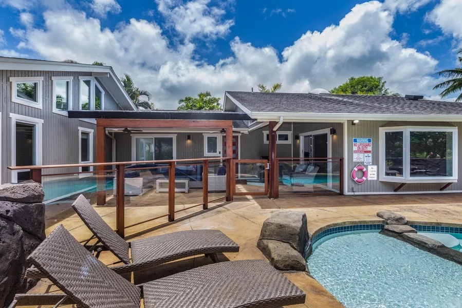 A backyard pool and patio sit beside a modern house under a cloudy sky.