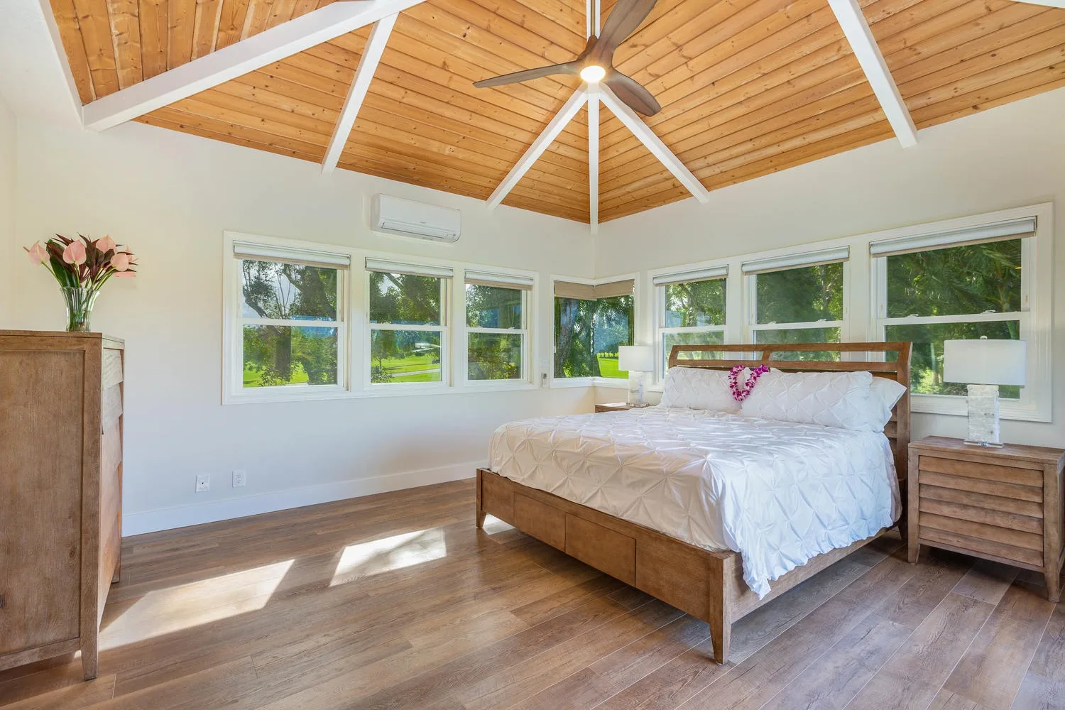 A bright bedroom with a wooden vaulted ceiling and windows overlooking greenery.