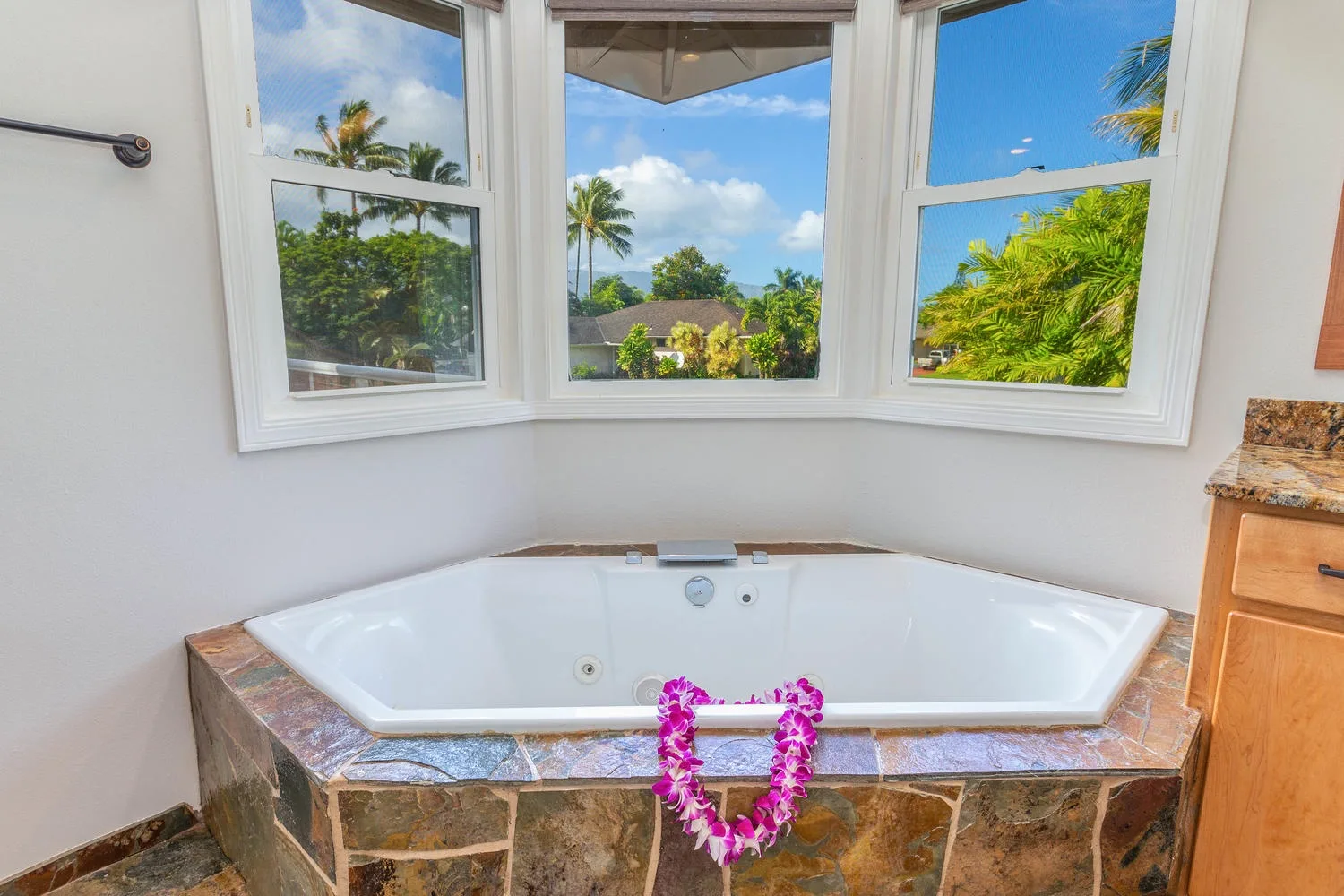 A bathtub sits beneath wide windows overlooking palm trees and greenery.