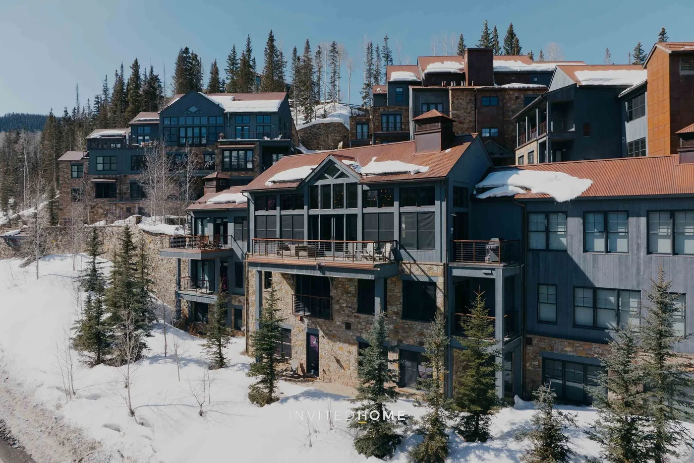 Snow-covered buildings sit among evergreen trees on a wooded hillside.